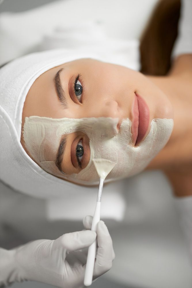 Woman receiving a facial treatment with a sheet mask being applied by a gloved practitioner.