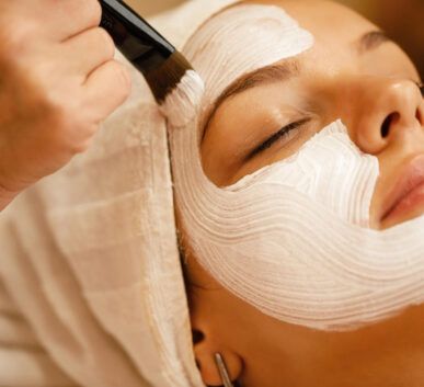 esthetician applies a thick white facial mask with a brush on a woman's face during a spa treatment.