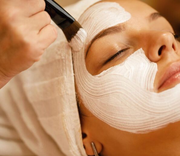 Higiene facial profunda esthetician applies a thick white facial mask with a brush on a woman's face during a spa treatment.