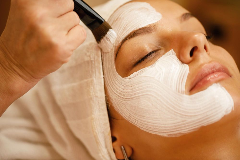 Esthetician applies a thick white facial mask with a brush on a woman's face during a spa treatment.