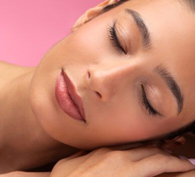 close-up of a woman with pink lipstick resting her head on her hands, eyes closed against a pink background.