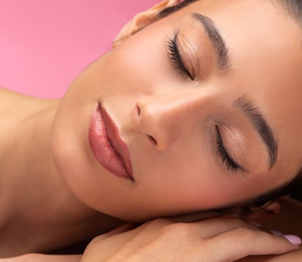 Microblading (3) close-up of a woman with pink lipstick resting her head on her hands, eyes closed against a pink background.