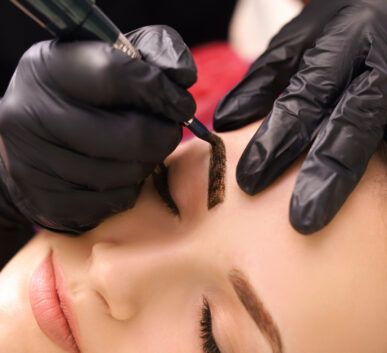 technician applying pigment to a woman's eyebrow during a microblading procedure with gloved hands.