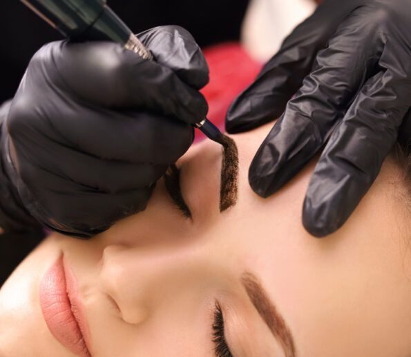technician applying pigment to a woman's eyebrow during a microblading procedure with gloved hands.