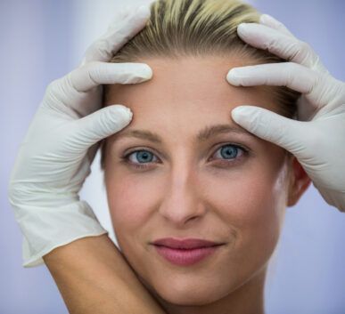close-up of a woman with blue eyes as gloved hands frame her face, suggesting a facial examination by a clinician.
