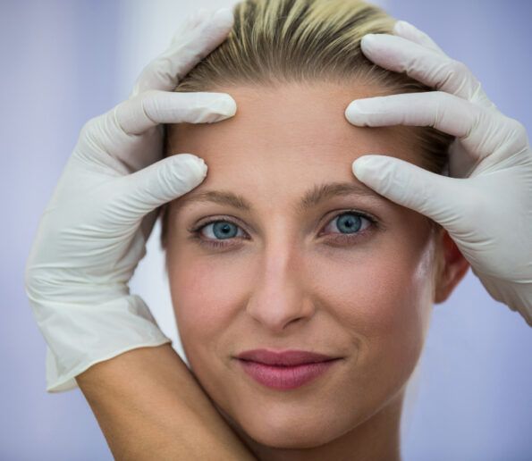 Tratamiento contorno de ojos close-up of a woman with blue eyes as gloved hands frame her face, suggesting a facial examination by a clinician.