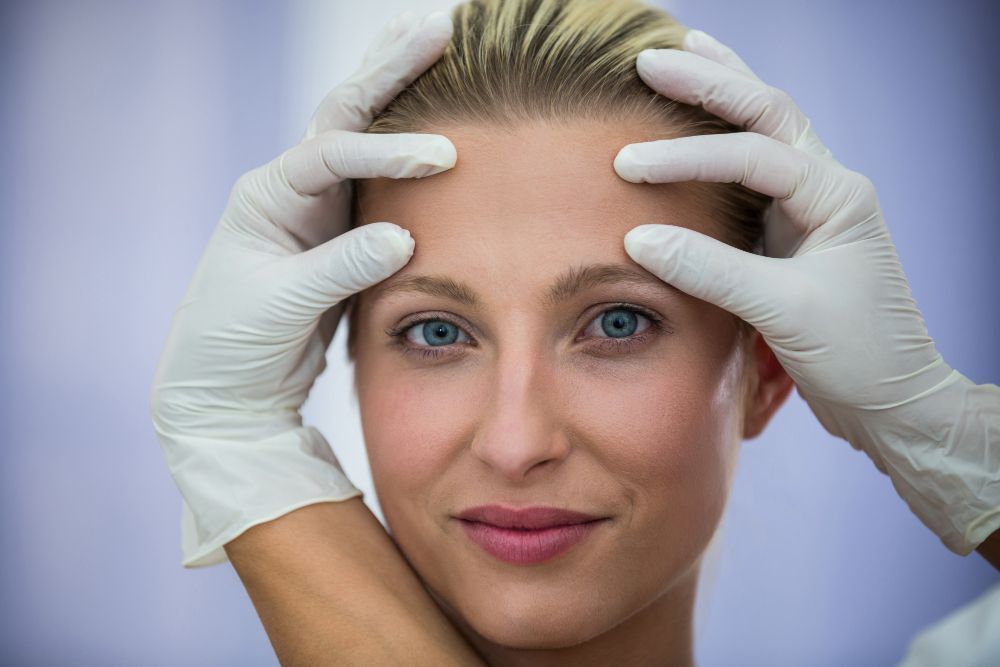 Close-up of a woman with blue eyes as gloved hands frame her face, suggesting a facial examination by a clinician.