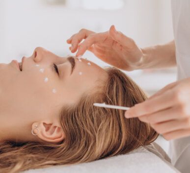 woman lying with eyes closed during a facial treatment as cream dots are applied to her face with a cotton swab by an esthetician.