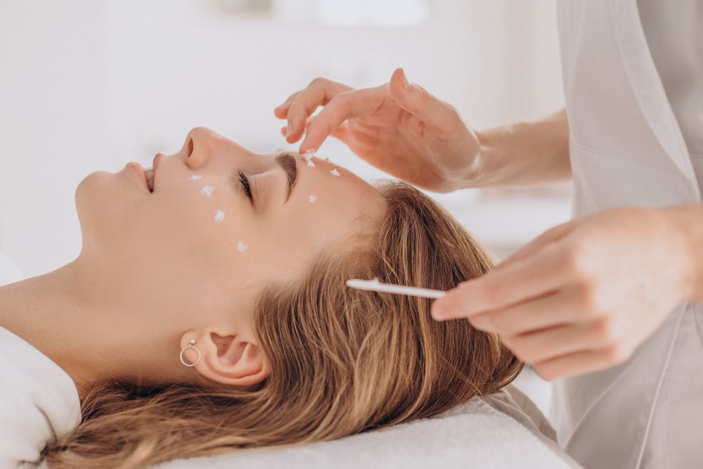 Woman lying with eyes closed during a facial treatment as cream dots are applied to her face with a cotton swab by an esthetician.