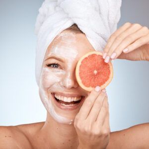 Woman with a white facial mask and towel wrapped around her head, smiling while holding a grapefruit slice over one eye in a spa-like setting
