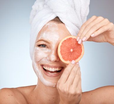 woman with a white facial mask and towel wrapped around her head, smiling while holding a grapefruit slice over one eye in a spa-like setting