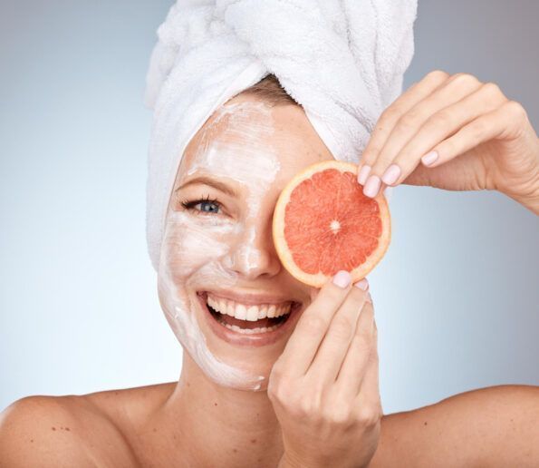 Tratamiento facial vitaminas y minerales woman with a white facial mask and towel wrapped around her head, smiling while holding a grapefruit slice over one eye in a spa-like setting