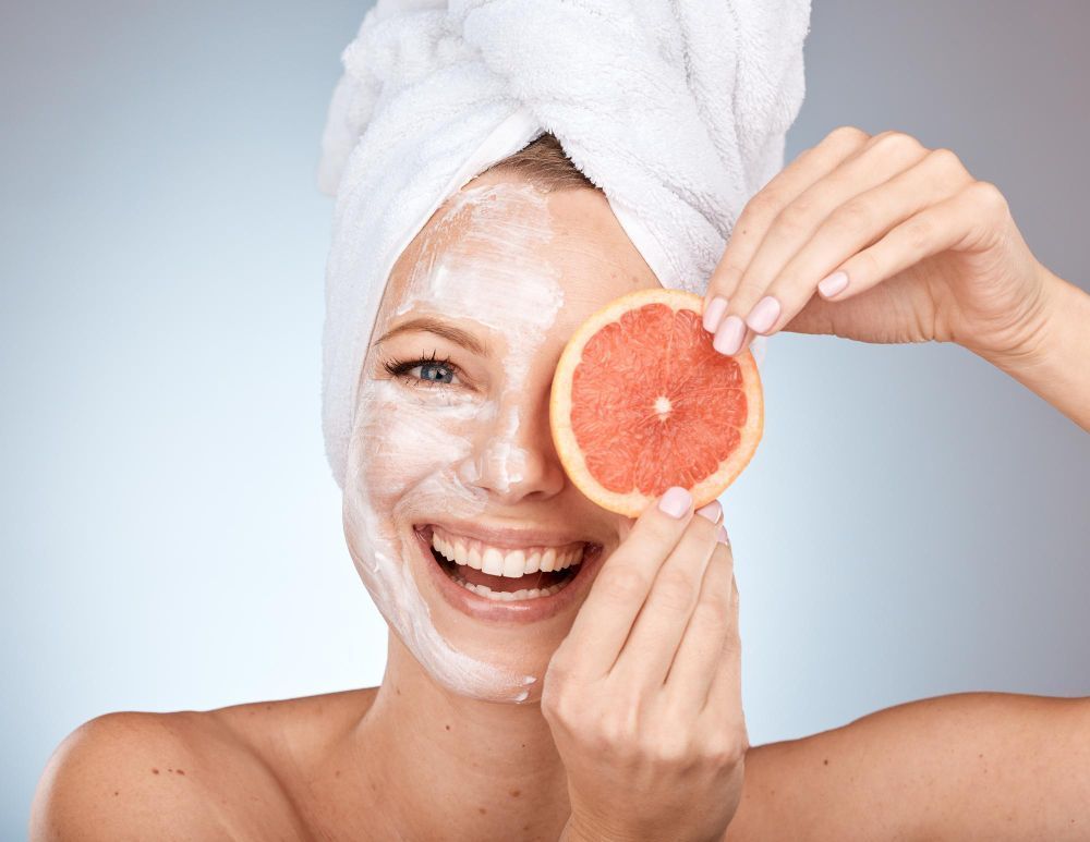 Woman with a white facial mask and towel wrapped around her head, smiling while holding a grapefruit slice over one eye in a spa-like setting