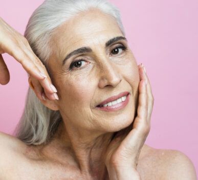 close-up of a smiling older woman with silver hair, hands framing her face against a pink background.