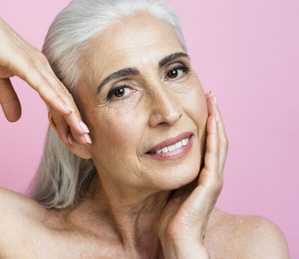 close-up of a smiling older woman with silver hair, hands framing her face against a pink background.