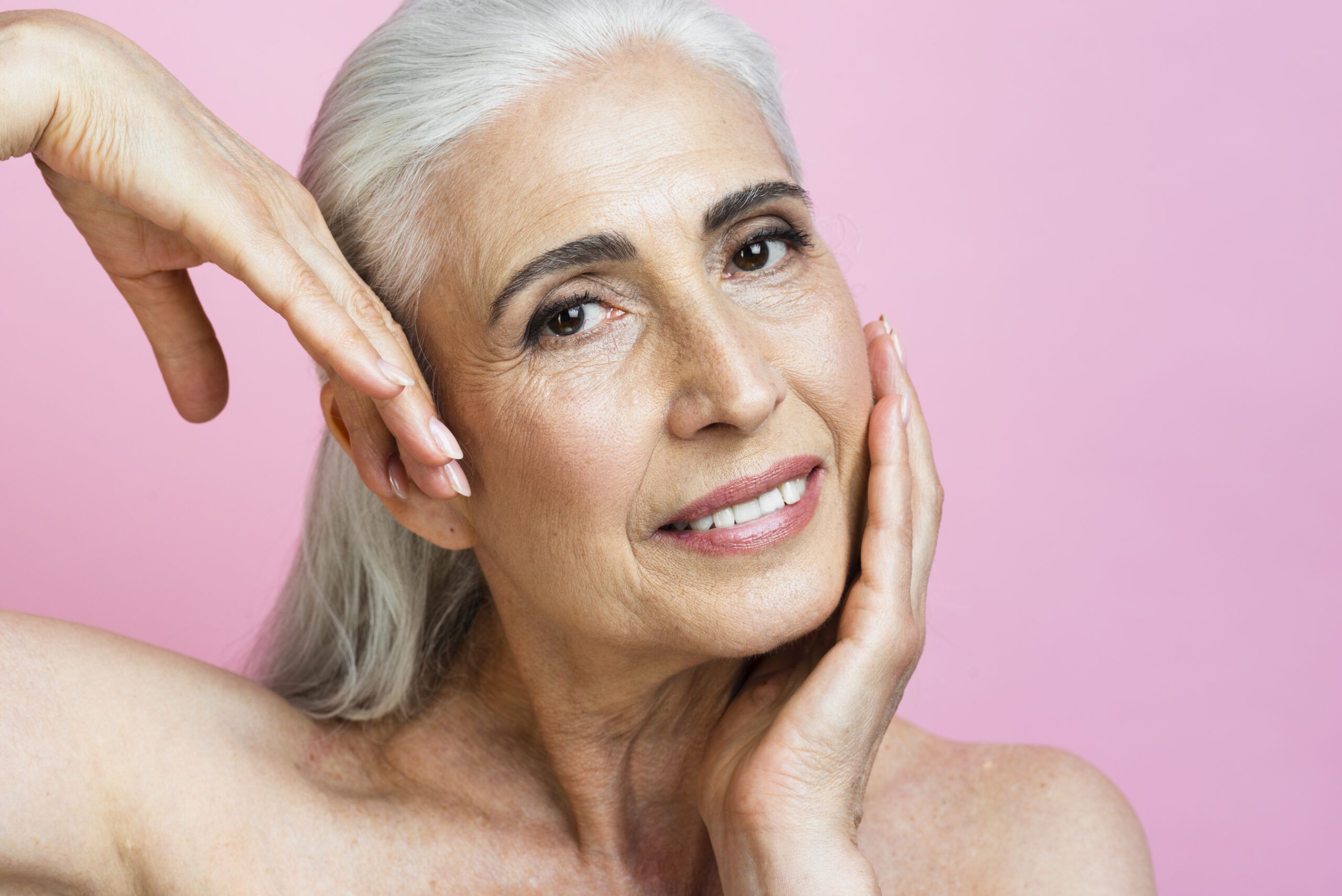 Close-up of a smiling older woman with silver hair, hands framing her face against a pink background.
