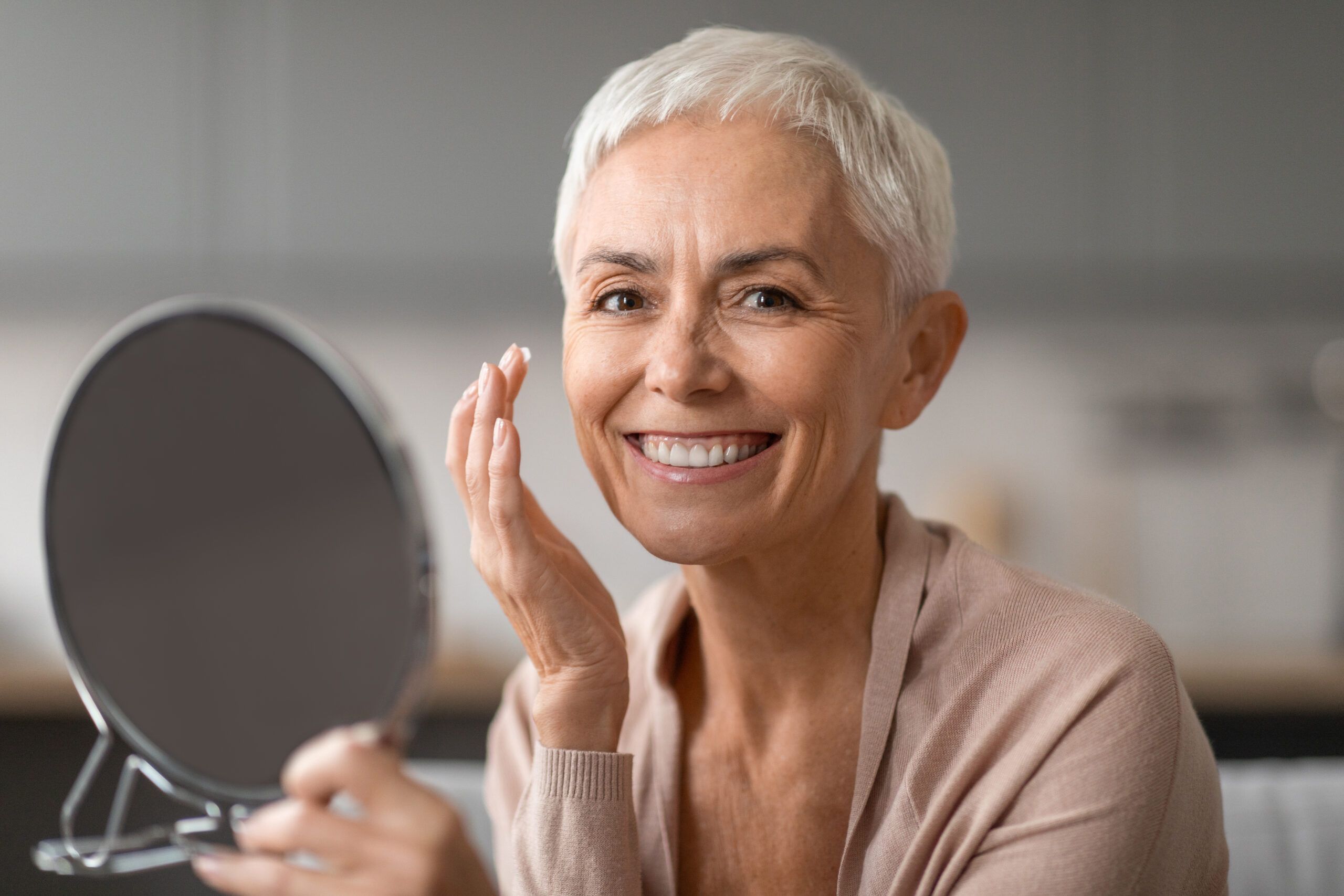 Senior woman with short white hair smiling as she looks into a mirror while applying makeup in a cozy setting.