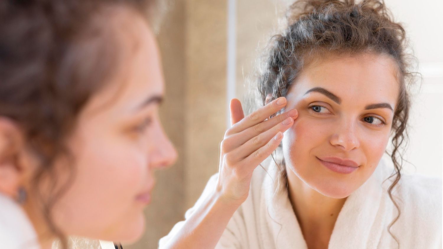 Woman with curly hair in a white robe touches the skin around her eye while looking at herself in a mirror, suggesting a skincare routine.