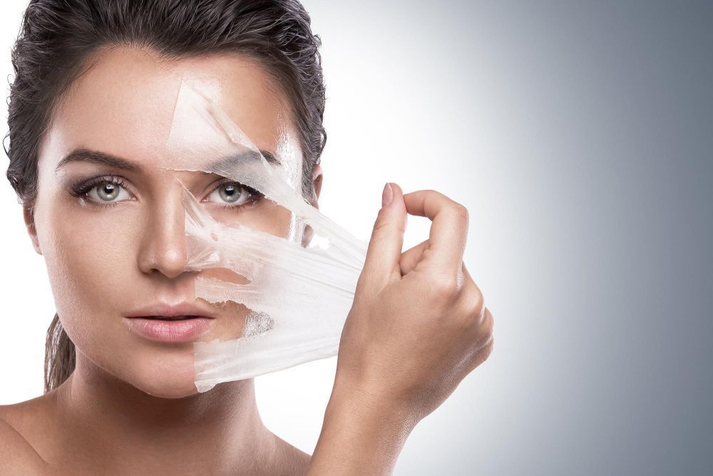 Woman peels off a clear facial sheet mask from half of her face, revealing smooth skin on the exposed side.