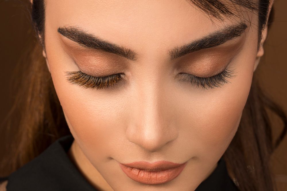 Close-up of a woman's face with eyes closed, wearing bronze eyeshadow, long lashes, bold brows, and peach lipstick.