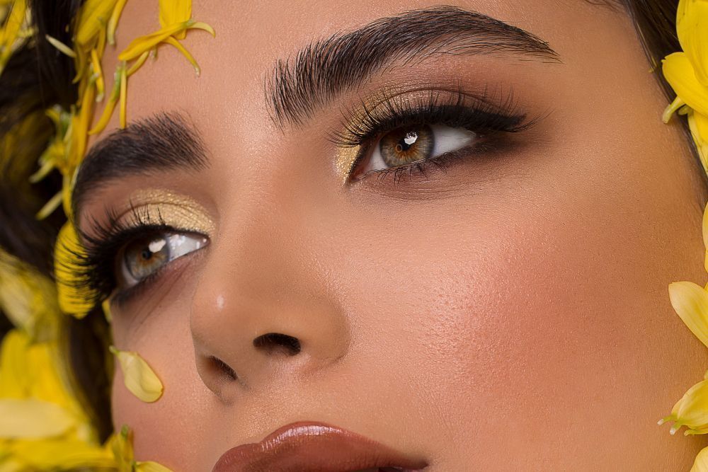 Close-up of a woman's face with gold eye makeup and yellow flower petals framing her features