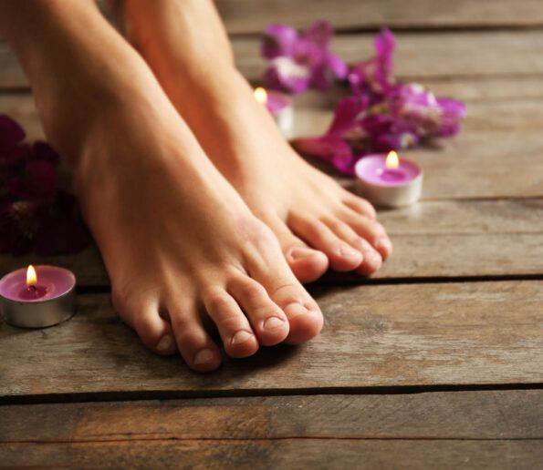 pedicura (5) pair of bare feet resting on a wooden floor surrounded by pink candles and purple flowers for a spa setting.