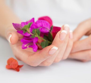 Close-up of hands with French-tipped nails holding a small bouquet of bright pink and purple flowers on a white surface, with an orange blossom nearby