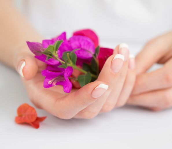close-up of hands with french-tipped nails holding a small bouquet of bright pink and purple flowers on a white surface, with an orange blossom nearby
