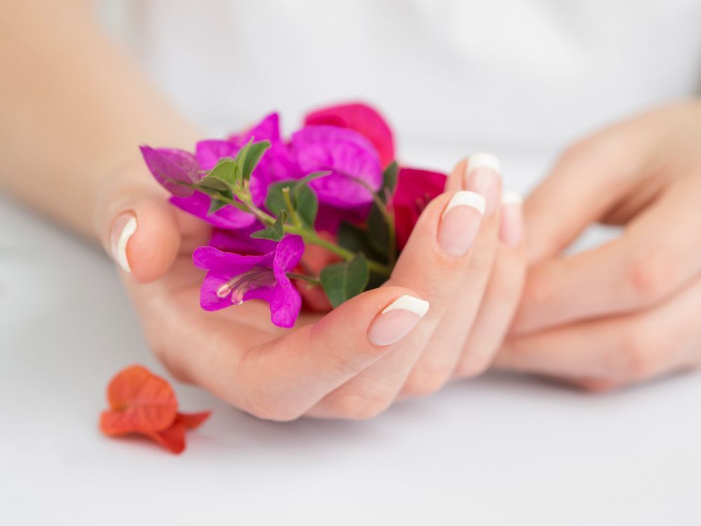 Close-up of hands with French-tipped nails holding a small bouquet of bright pink and purple flowers on a white surface, with an orange blossom nearby